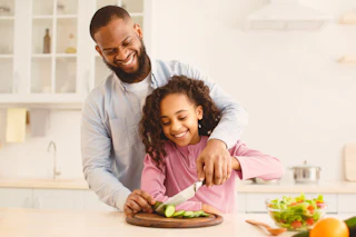 Helper. Portrait of cheerful black father teaching his daughter how to cook, holding knife together while cutting vegetables, slicing cucumber, little girl learning how to prepare healthy fresh salad