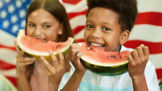 Portrait of smiling African boy holding watermelon and looking at camera with American flag in background, copy space