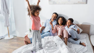 excited african american girl standing on bed with raised hands near smiling parents and brother