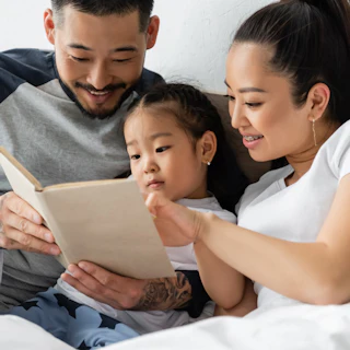 smiling asian parents reading book to toddler daughter in bed